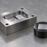 Machined steel tooling block beside a black mount holding a steel specimen