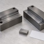 Two stepped steel tooling blocks beside a small steel coupon and blank card